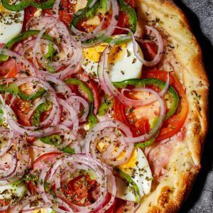 Close-up of a colorful vegetable pizza topped with sliced eggs and herbs on a stone surface.